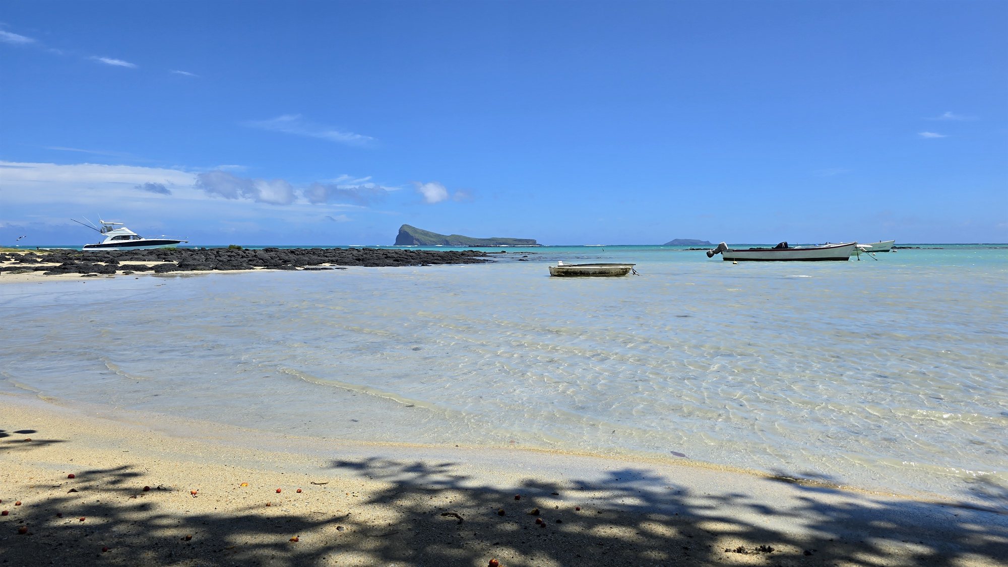 Sunlit Mauritius coast with boats in shallow water and palm shadows on the shore.
